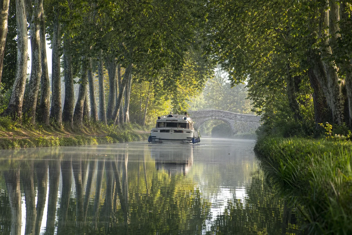 Coche de plaisance progressant lentement entre deux berges plantees, eau calme et reflets du matin